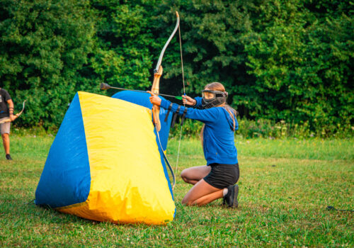 Junge Frau kniet beim Arrow Tag hinter einem aufblasbaren Schutz, um eine gute Position zum Abschießen ihres Pfeils zu bekommen