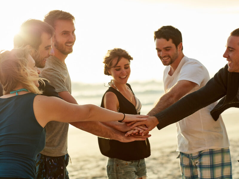 Freunde halten in einem Kreis ihre Hände zusammen für ein Teambuilding am Strand.