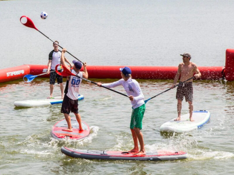 Vier Spieler auf SUP-Boards spielen in einem abgegrenzten Bereich eines Sees SUP Polo.