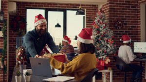 Büroangestellte tauschen bei der Arbeit Weihnachtsgeschenke aus und fühlen sich festlich mit Weihnachtsmannmützen im Büro mit Weihnachtsbaum und Dekorationen. 